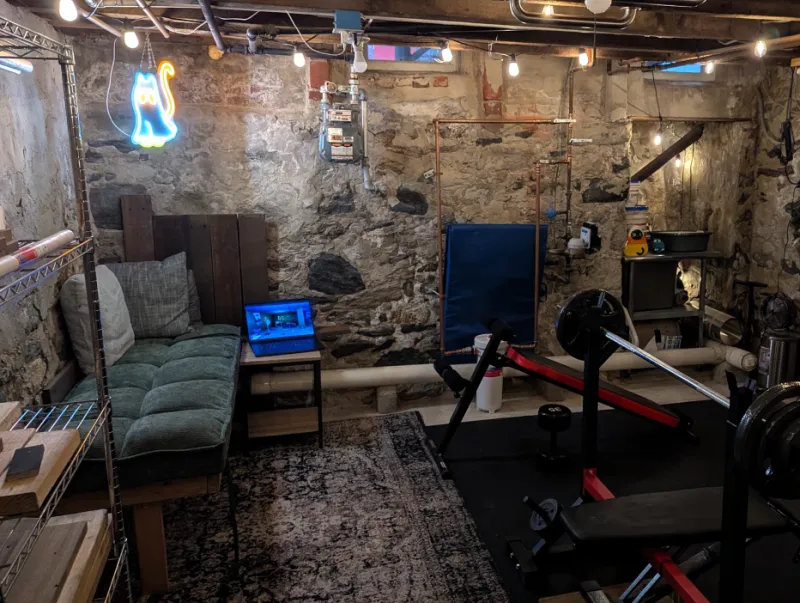Wide view of the basement nook with the daybed, a laptop on a side table, a rug, and a weight bench